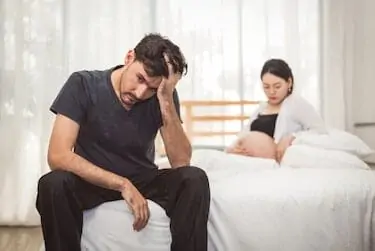 Worried stress man sitting on bed in serious mood emotion with pregnant wife woman background