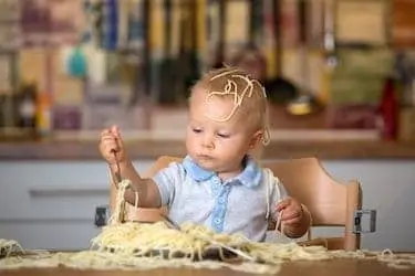 Little baby eating spaghetti for lunch and making a mess at home in kitchen