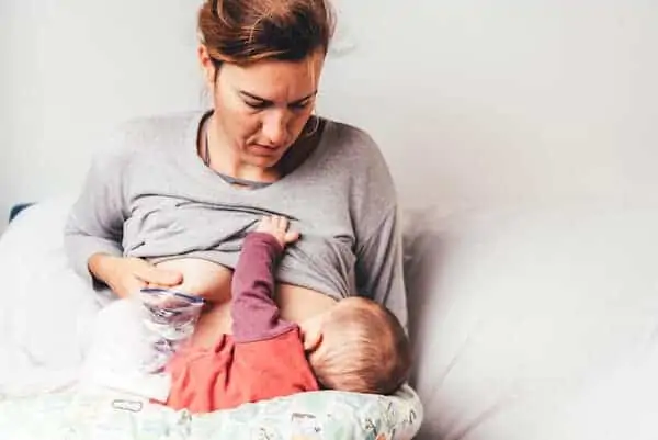 Mother breastfeeding newborn while pumping milk to store it in a bag